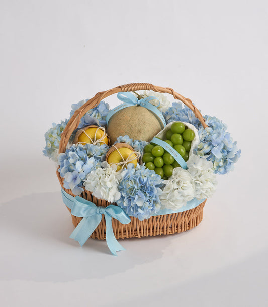 Wicker basket with blue and white hydrangeas, featuring sliced lemons, green grapes, and a tan sphere, tied with light blue ribbon.