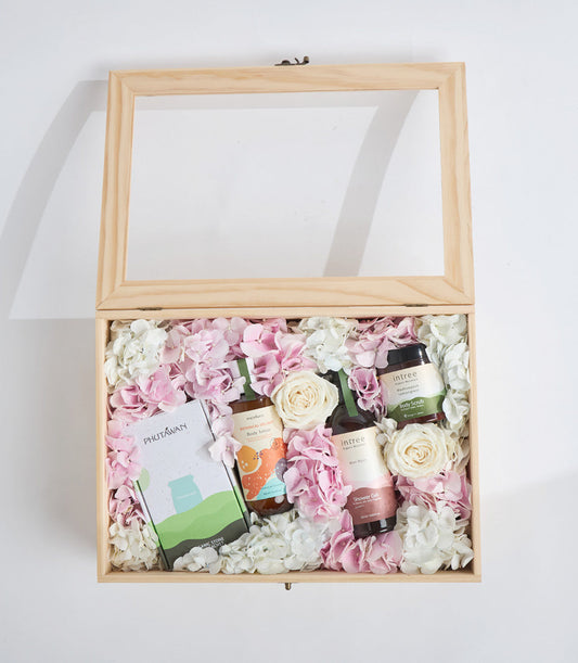 Luxury spa gift box with glass lid, natural wood frame, pink and white flowers, skincare products, and beauty items arranged on white background.