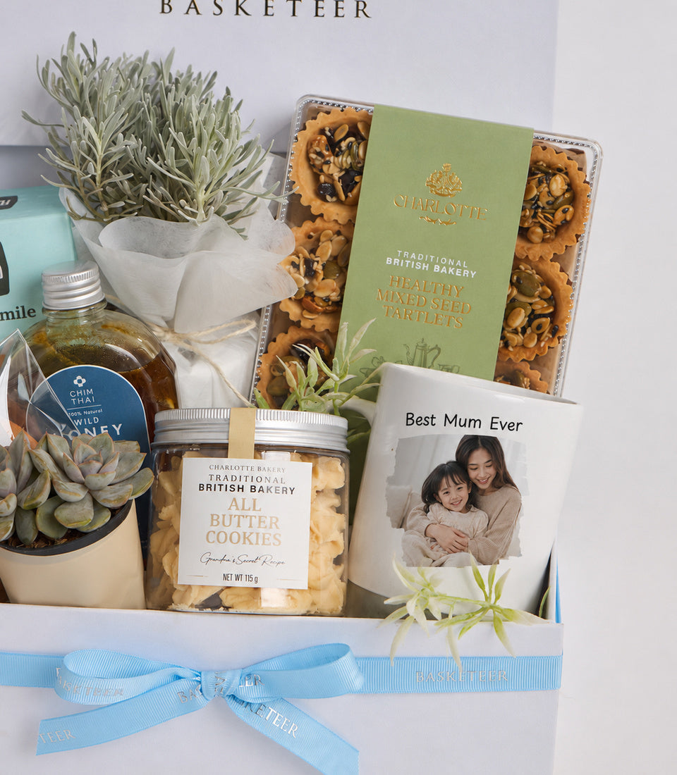 A white "BASKETEER" gift basket containing Charlotte Bakery treats, succulents, honey, a "Best Mum Ever" mug, and blue ribbon bow.