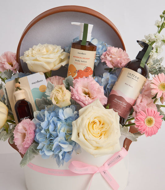 Luxury spa gift basket with brown bottles, fresh flowers in cream, pink and blue tones, wrapped in white box with pink ribbon.