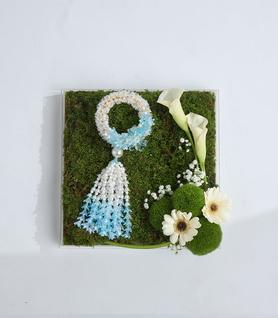 Decorative moss wall art featuring a blue and white beaded ornament with tassel, white calla lilies, and baby's breath on green moss background.