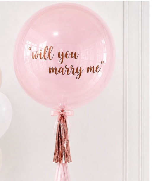 # Alt Text
Large pink clear balloon with "will you marry me" text in rose gold, decorated with rose gold and pink tassels.