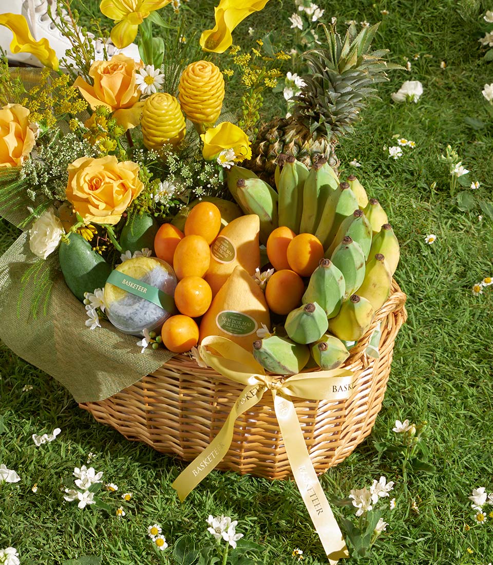 Woven wicker fruit basket overflowing with tropical mangoes, bananas, pineapple, and tangerines, adorned with yellow roses