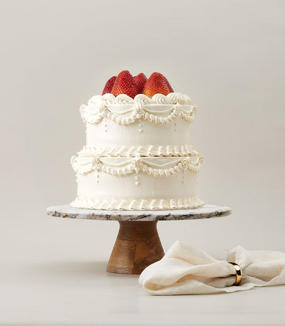 Two-tier strawberry cream cake with white frosting, pearl beads, and fresh strawberries on marble stand