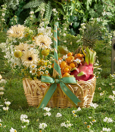 Wicker basket with tropical fruits, cream gerbera daisies, and white flowers tied with green gold-edged ribbon on garden lawn