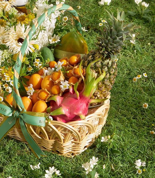 Woven wicker basket with tropical fruits, fresh flowers and green-gold ribbon on grass