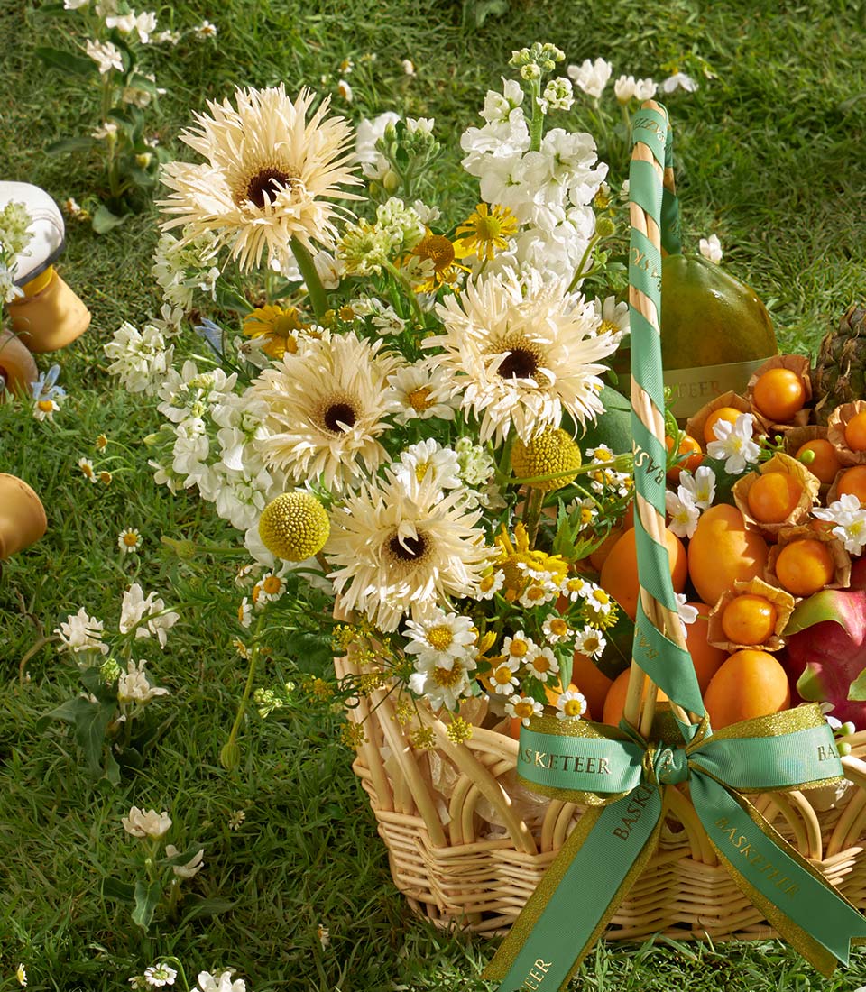 Light yellow and white gerbera daisies with fresh tropical fruits in a premium wicker basket gift