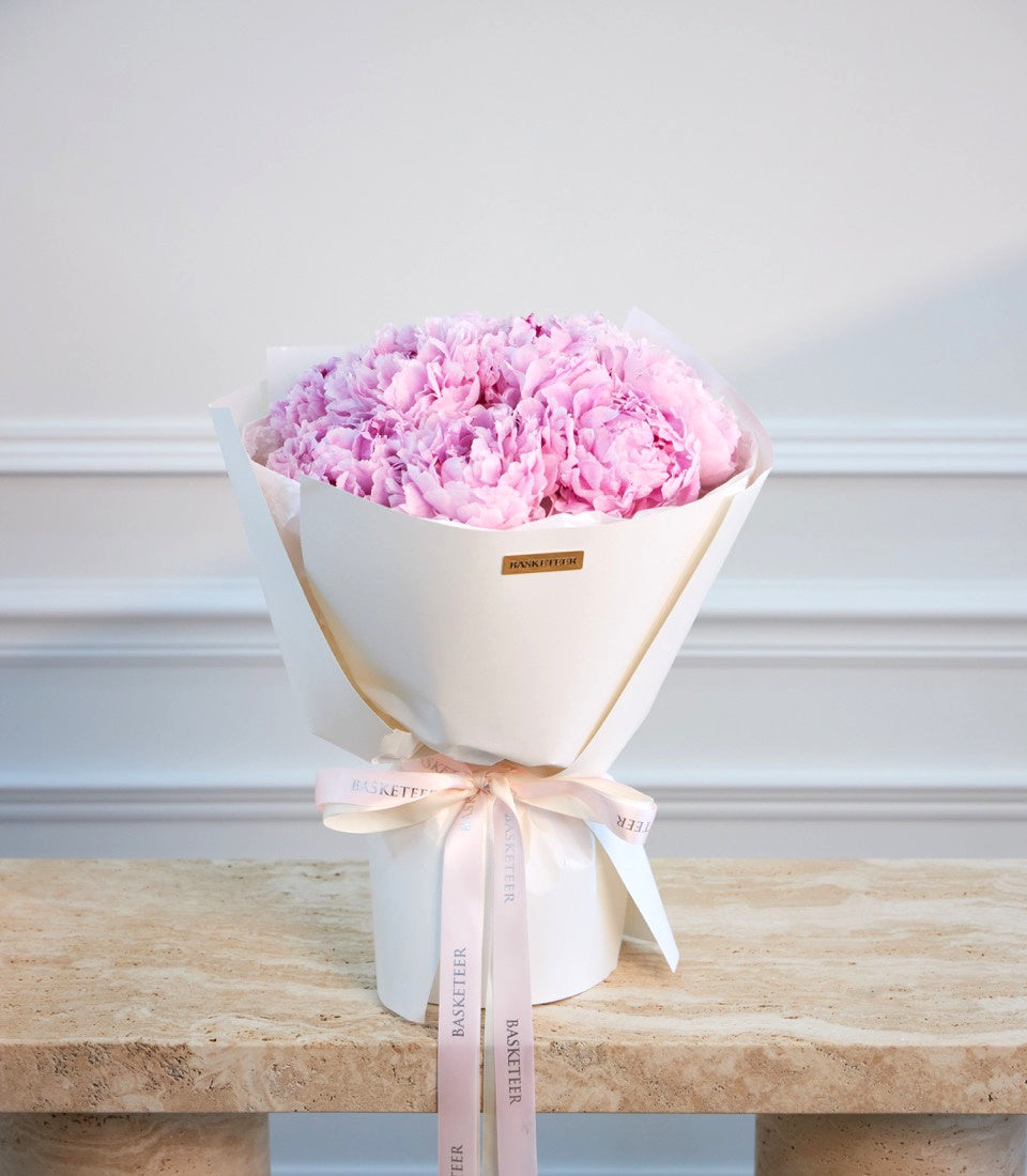 Luxurious bouquet of pink peonies in white paper wrapper with pink ribbon bow, branded "Basketeer" label, displayed on marble surface.