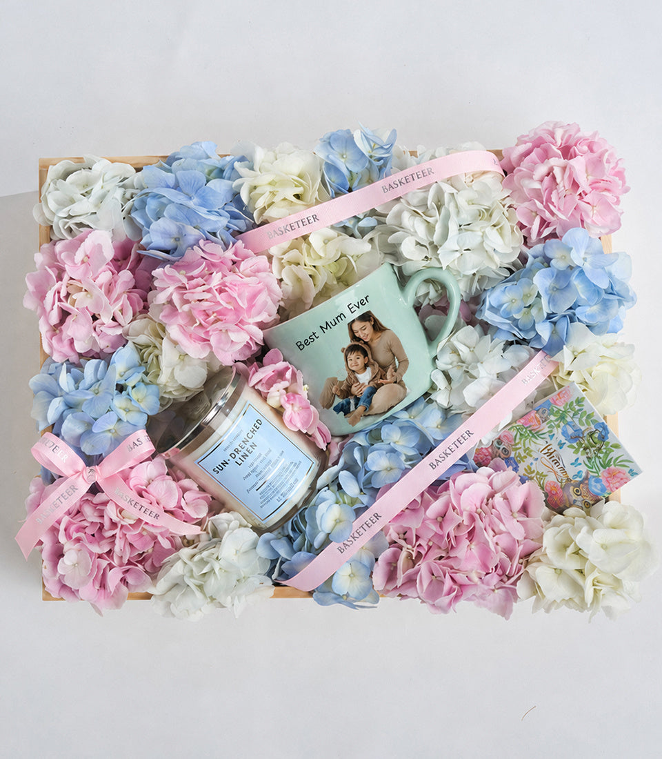 Luxe gift basket with pink, blue, and cream hydrangeas, personalized photo card, candle, and Basketeer branded items on wooden base.