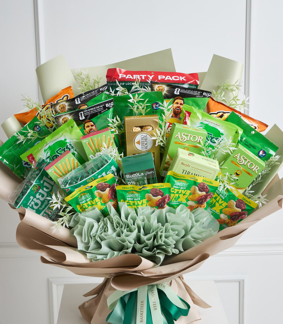 Colorful gift bouquet with green and orange snack packages, decorative white and sage tissue paper, dried grass accents, and beige wrapping, displayed against white wall background.