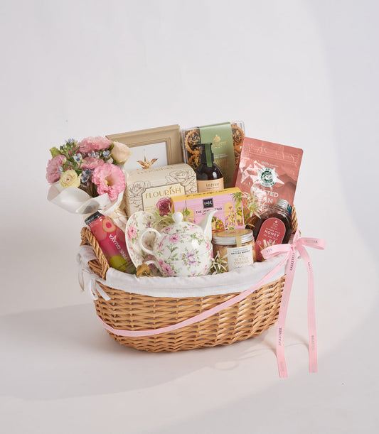 Luxury wicker gift basket with pink flowers, floral teapot, gourmet treats, wine, and decorative items tied with pink ribbons.