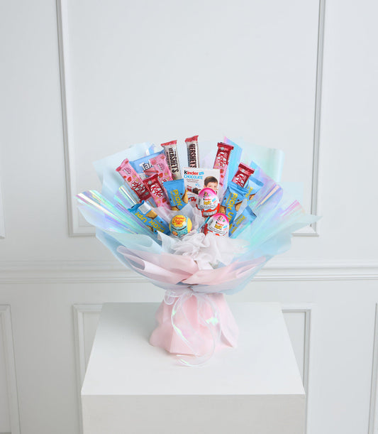 Colorful candy bouquet with pastel pink and blue cellophane wrapping, featuring Hershey's, Kinder, and Lollipop treats arranged in a fan display on white cube pedestal.