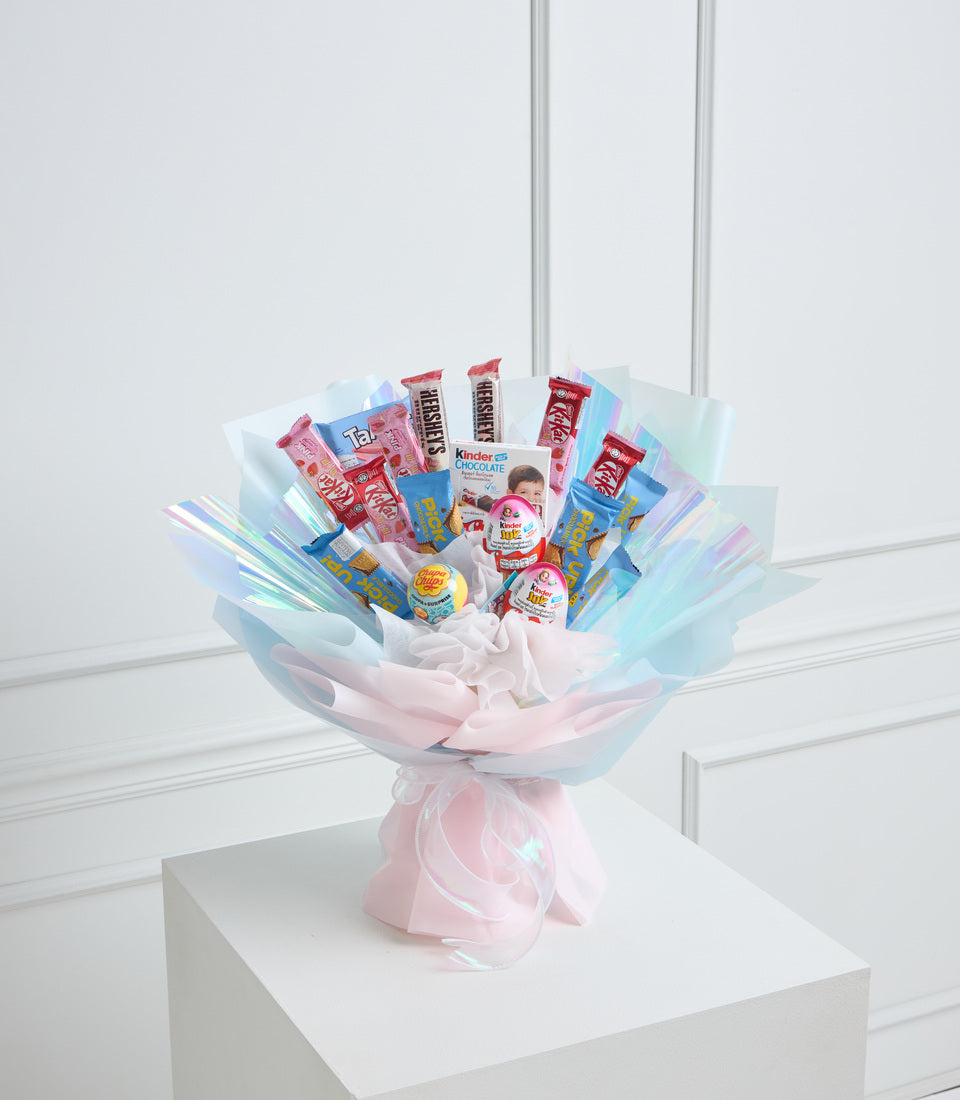 Candy bouquet with pink and blue cellophane wrapping, featuring chocolate bars, Kinder products, and sweets arranged in a decorative display on a white pedestal.