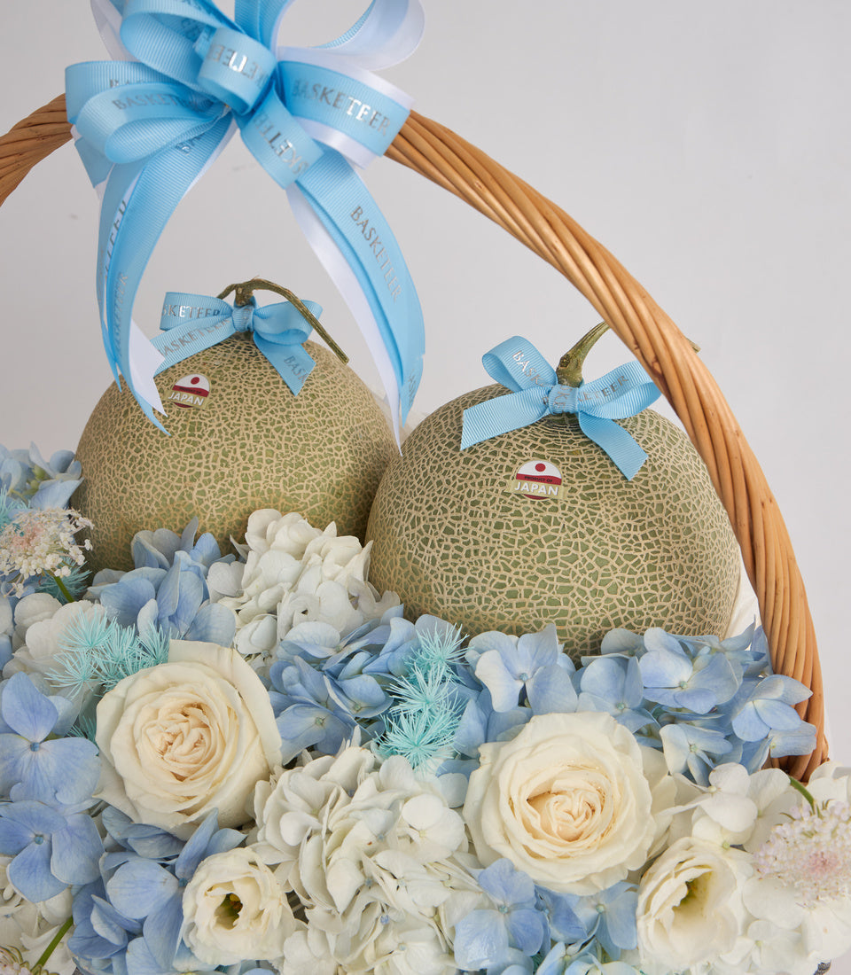 Luxury gift basket with two Japanese melons adorned with blue ribbons, surrounded by blue hydrangeas, white roses, and baby's breath flowers.