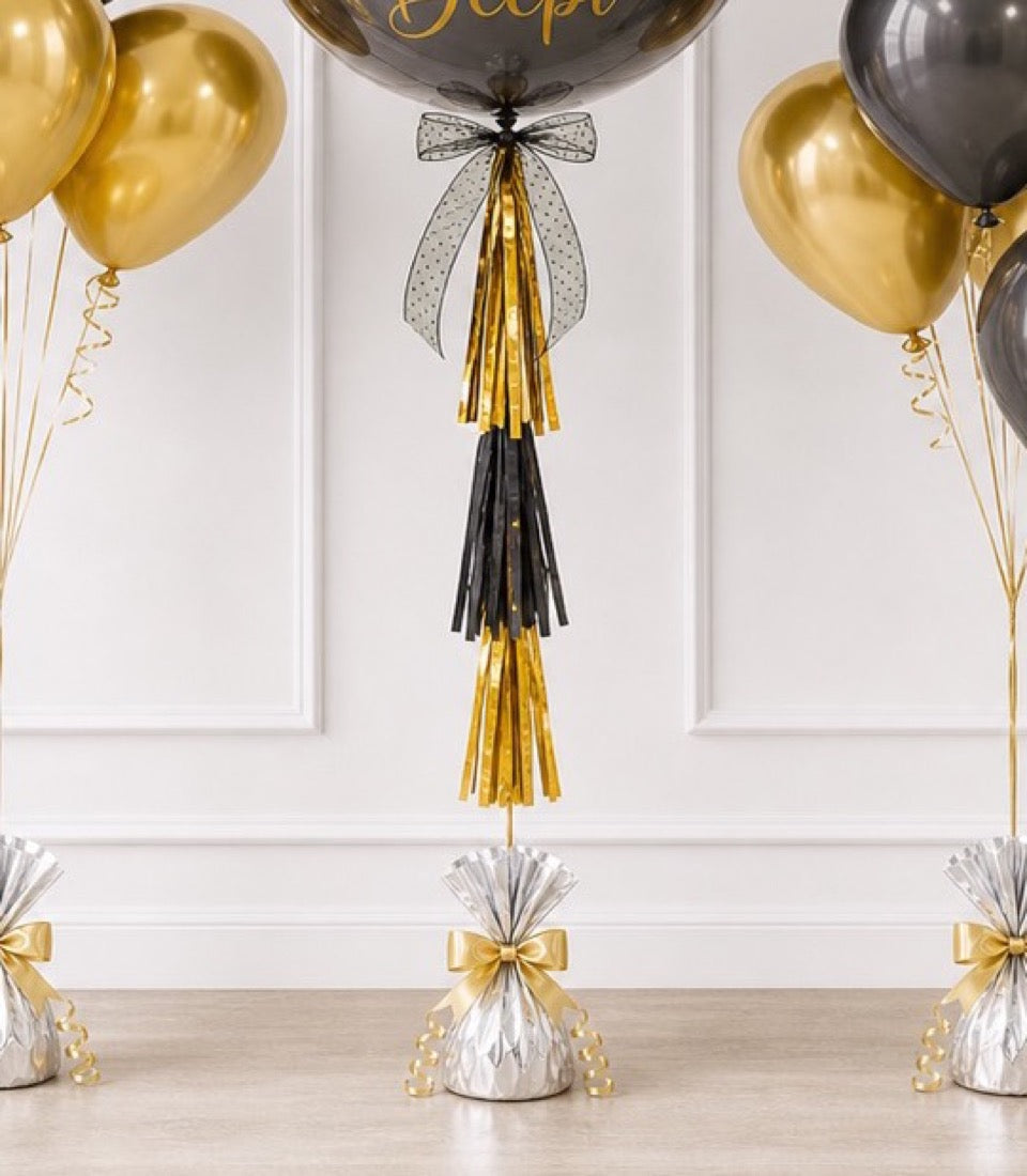 Elegant party balloon decoration with gold and black tassel garland, polka dot bow, and matching balloons in gold and silver on white paneled wall.