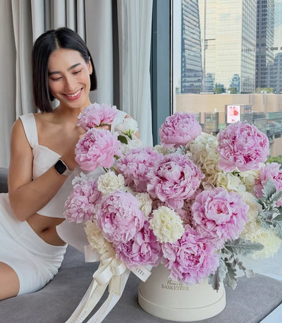 # Alt Text
Woman admiring luxurious arrangement of pink and white peonies in cream cylindrical vase with ribbon, high-rise city view behind.