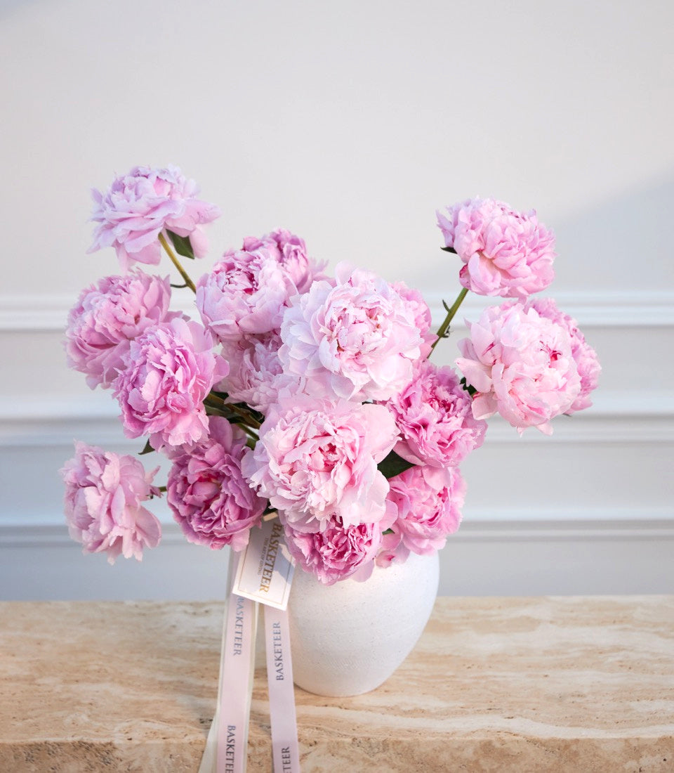 Pink peonies in a white ceramic vase with a pink ribbon bow on a wooden surface against a white wall.