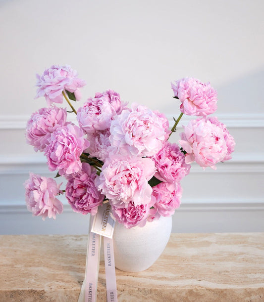 Pink peonies in a white ceramic vase with a pink ribbon bow on a wooden surface against a white wall.