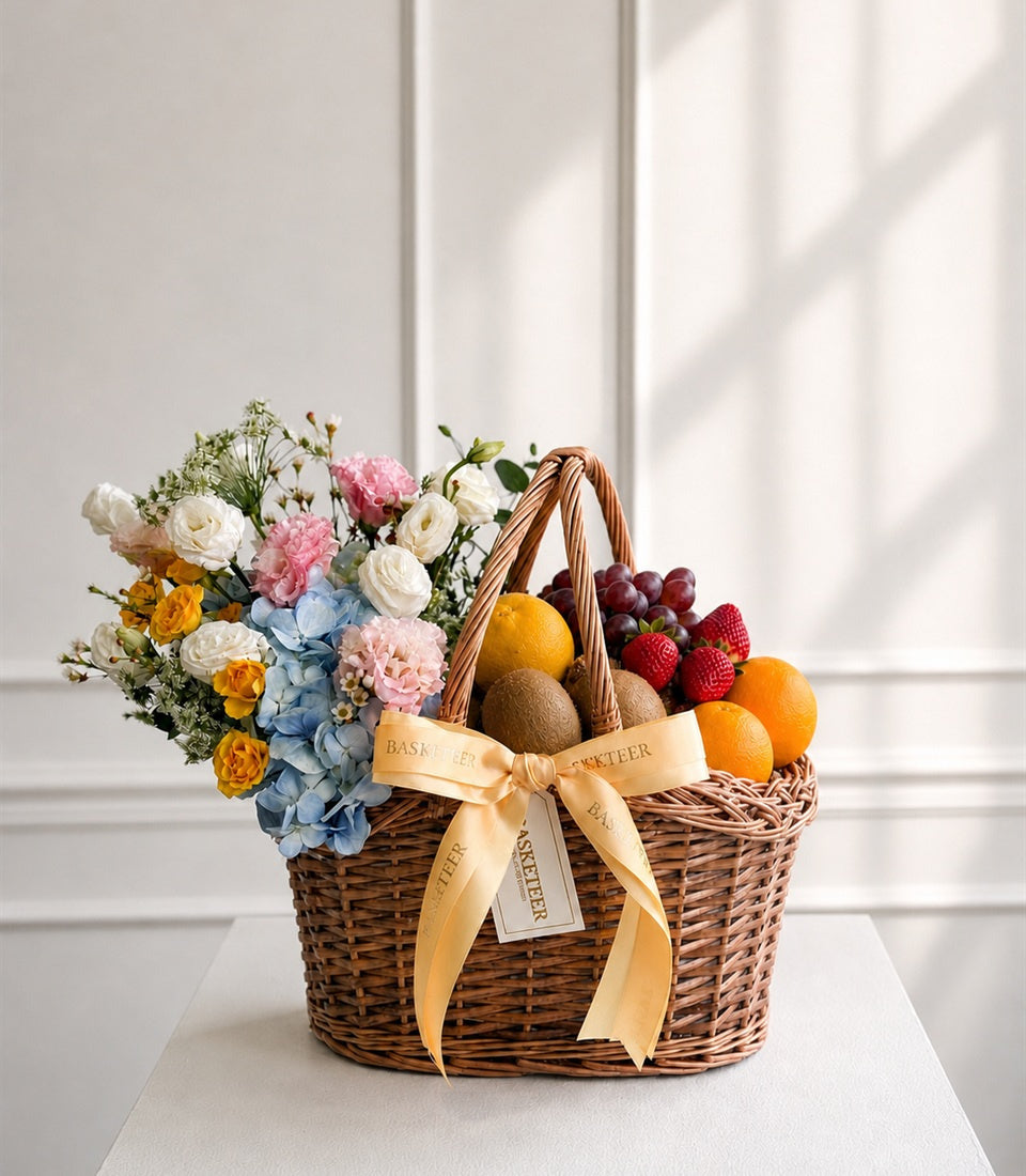 Wicker basket with fresh flowers, fruits, and gold ribbon bow against white background. Contains roses, hydrangeas, berries, citrus, and kiwis.