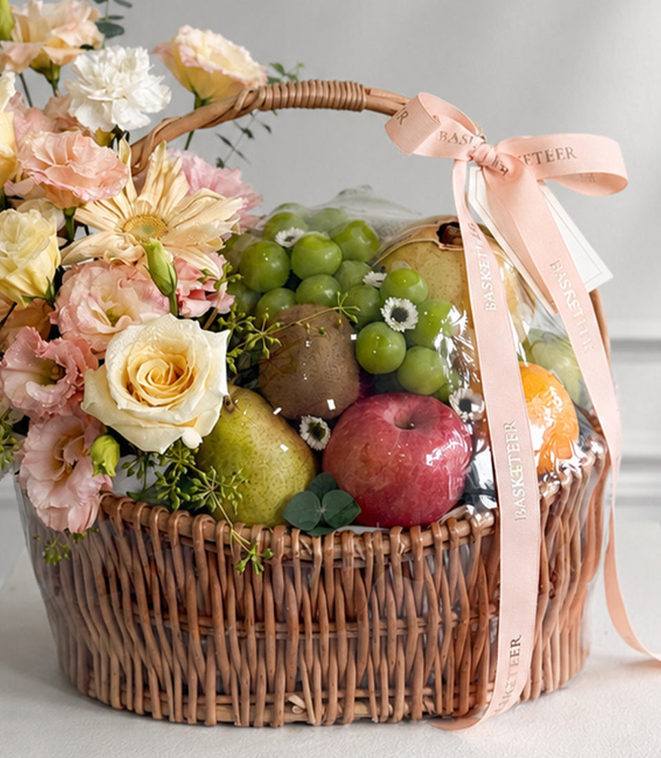 Wicker basket with fresh flowers, fruits, and a pink ribbon bow, featuring roses, daisies, pears, apples, and grapes in warm, elegant style.