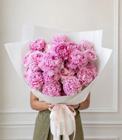Luxurious bouquet of pink peonies wrapped in white paper with pale pink ribbon, held by person in olive green shirt.