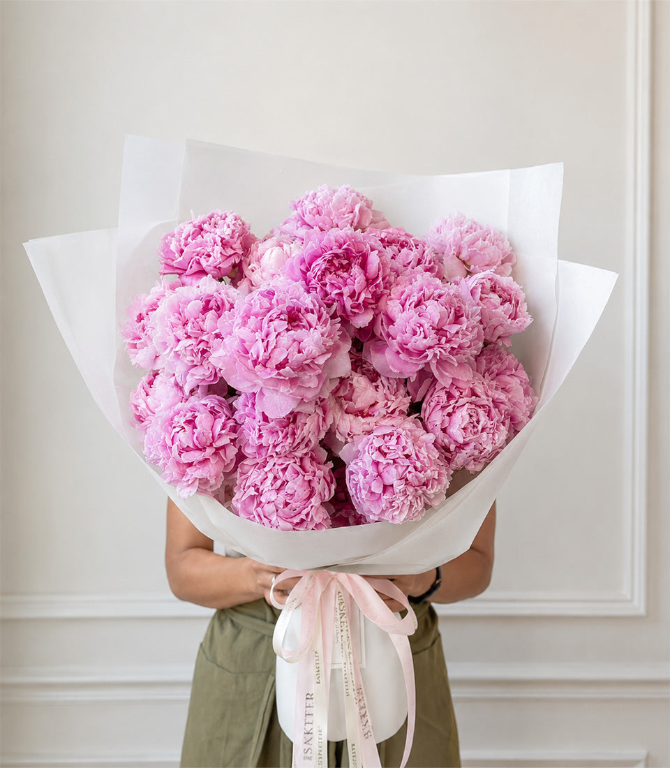 Luxurious bouquet of pink peonies wrapped in white paper with pale pink ribbon, held by person in olive green shirt.