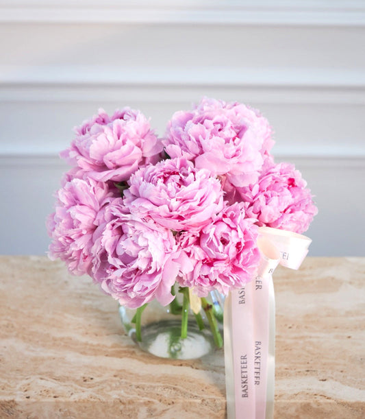 Pink peonies in clear glass vase with green stems, tied with pale pink ribbon labeled "Basketeer," on wooden surface.