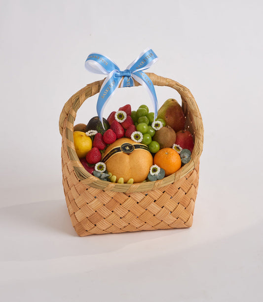 Woven wicker fruit basket with blue ribbon bow, containing raspberries, grapes, pears, oranges, kiwis, blueberries and decorative daisies.