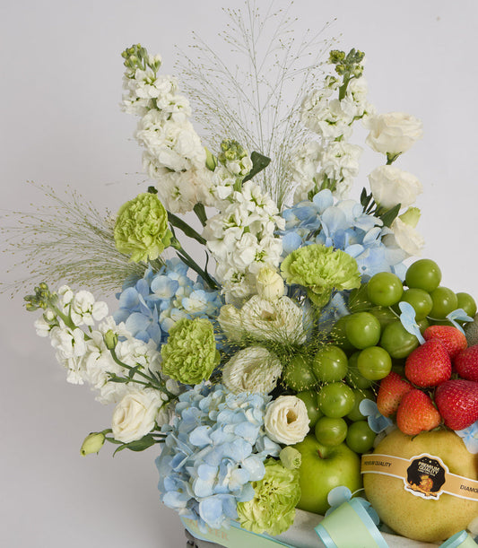 Luxurious floral and fruit arrangement featuring white stocks, blue hydrangeas, green chrysanthemums, ornamental grasses, fresh strawberries, green grapes, and pears on white background.