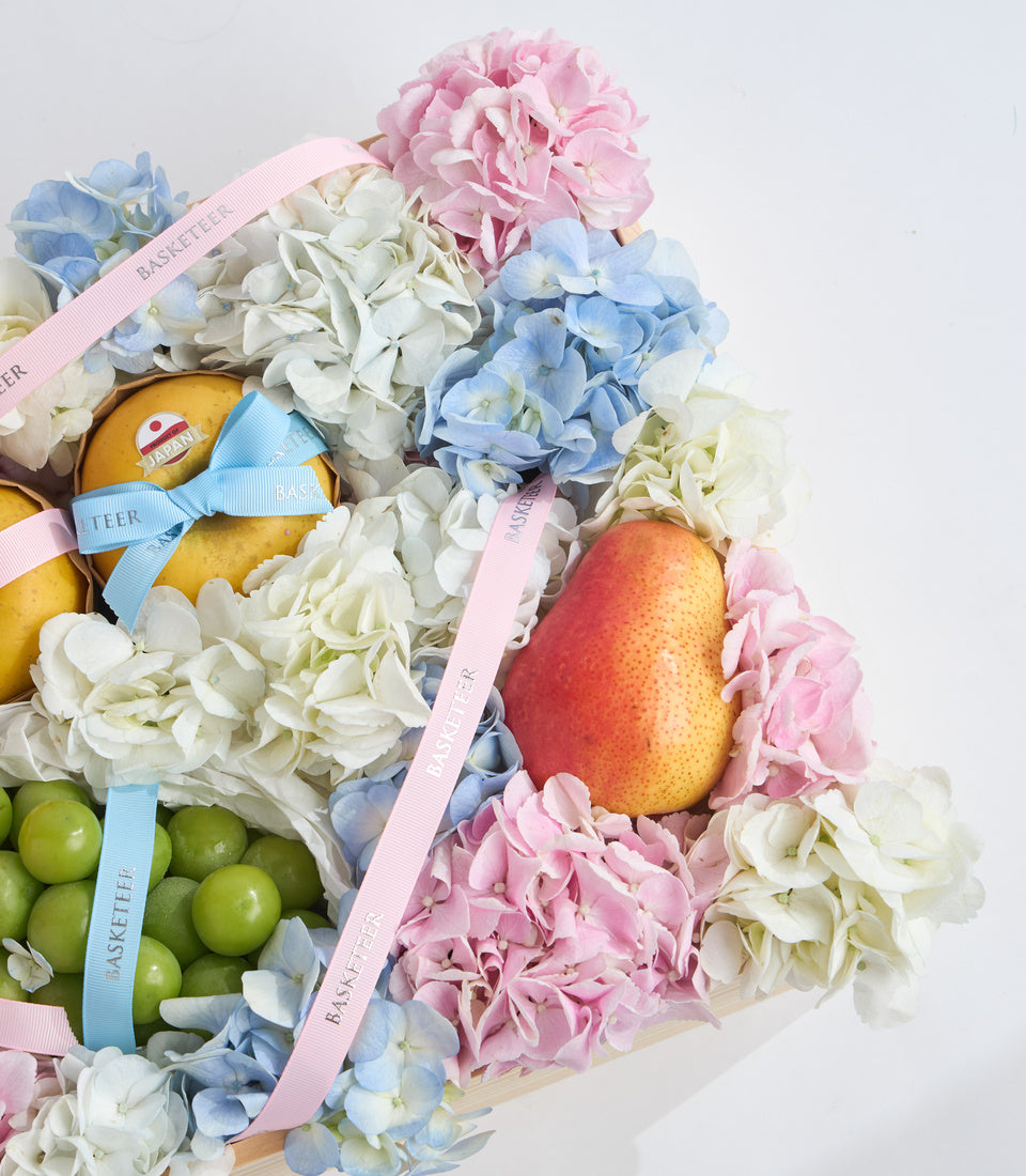 Luxury fruit and flower gift basket with pink peonies, blue and white hydrangeas, yellow lemons, green apples, and red mango tied with pastel ribbons.