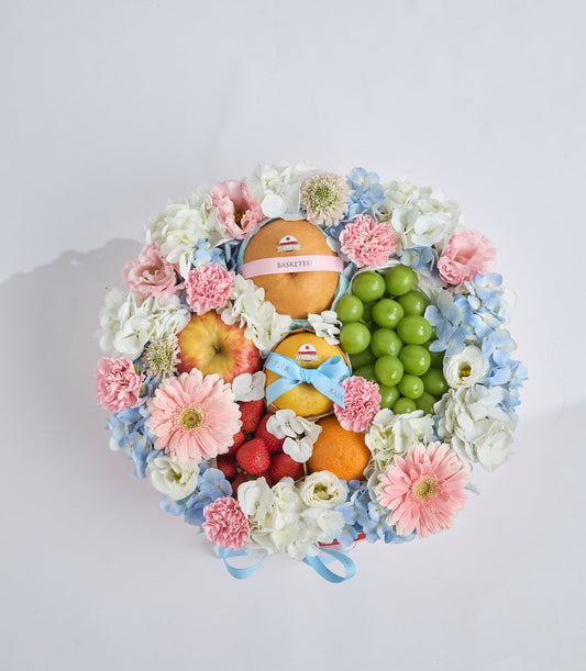 Floral gift basket with pink carnations, white roses, blue hydrangeas, fresh fruits including apples, oranges, green grapes, and raspberries, tied with blue and pink ribbons.