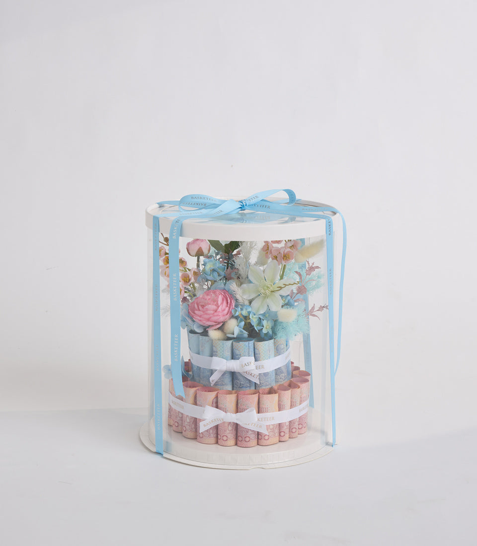 Cylindrical gift box with blue ribbon bow containing tiered arrangement of pink and blue rolled towels with fresh flowers including pink roses and white blooms.