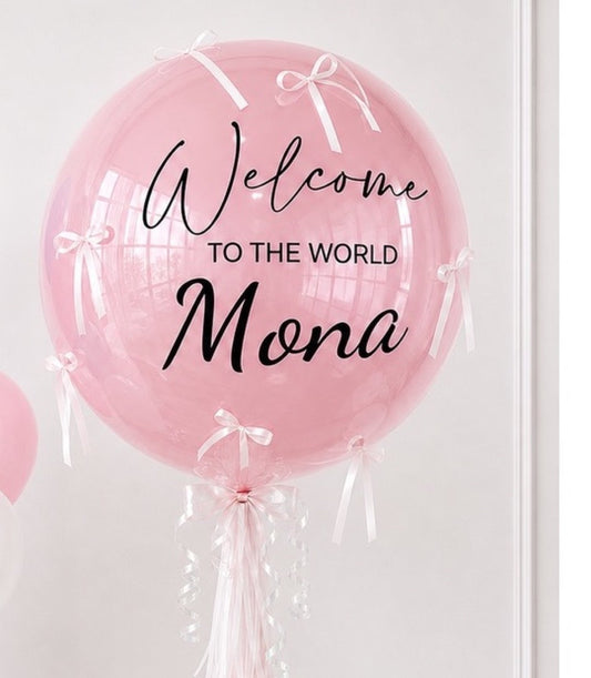 Pink bubble balloon with white ribbons and bows, printed text reading "Welcome to the World Mona" in black script and sans-serif fonts.