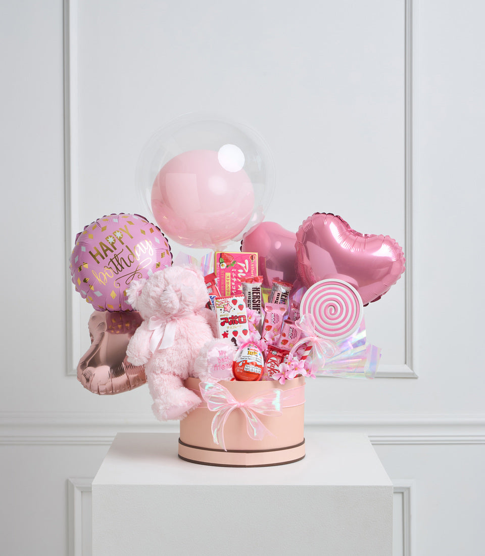 Pink birthday gift basket with balloons, plush teddy bear, candies, lollipop, and decorative elements in a cream-colored box.