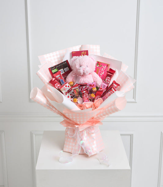Pink and white checkered bouquet with plush teddy bear, assorted chocolates, candies, and red gift boxes on white pedestal.