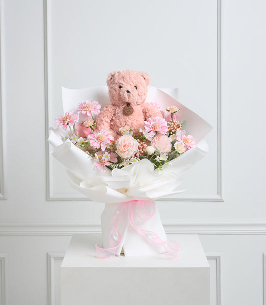 Pink teddy bear sitting in a bouquet of pink and white flowers with white wrapping and pink ribbon bow, displayed on white pedestal.