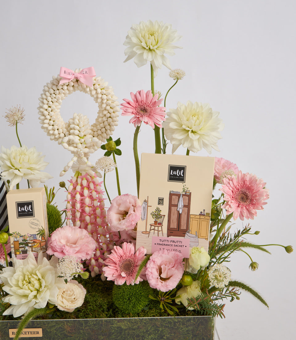 Elegant floral arrangement with cream and pink flowers, pearl beaded wreath, fragrance sachets, and moss in a dark container against white background.