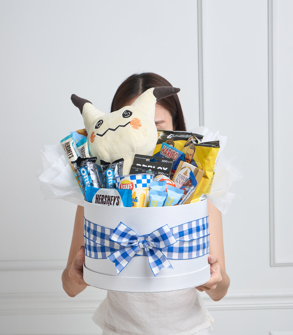 Woman holding white cylindrical gift basket with blue gingham bow, filled with snacks and a cream-colored plush cat toy with dark ears.
