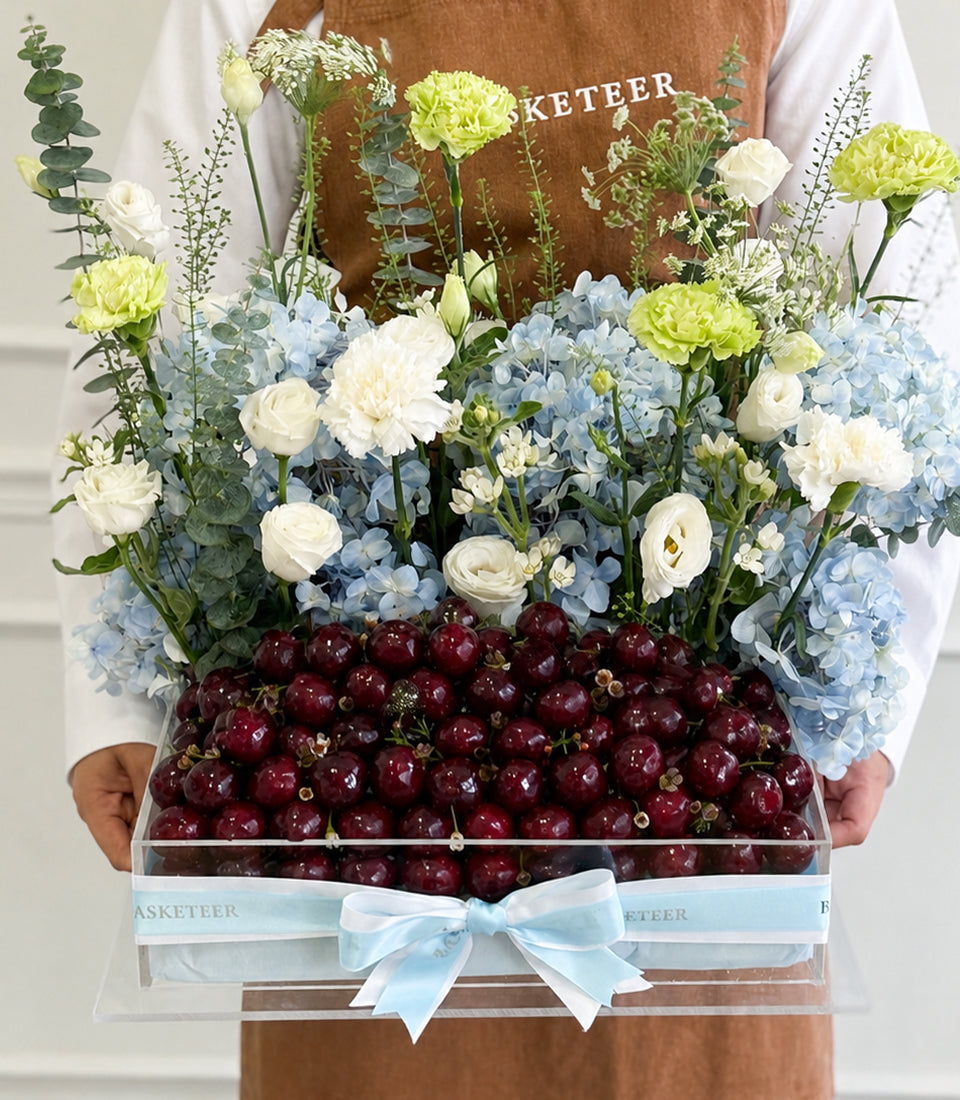 Florist holds luxury gift box with fresh cherries, white and green carnations, blue hydrangeas, white roses, greenery and light blue ribbon bow.