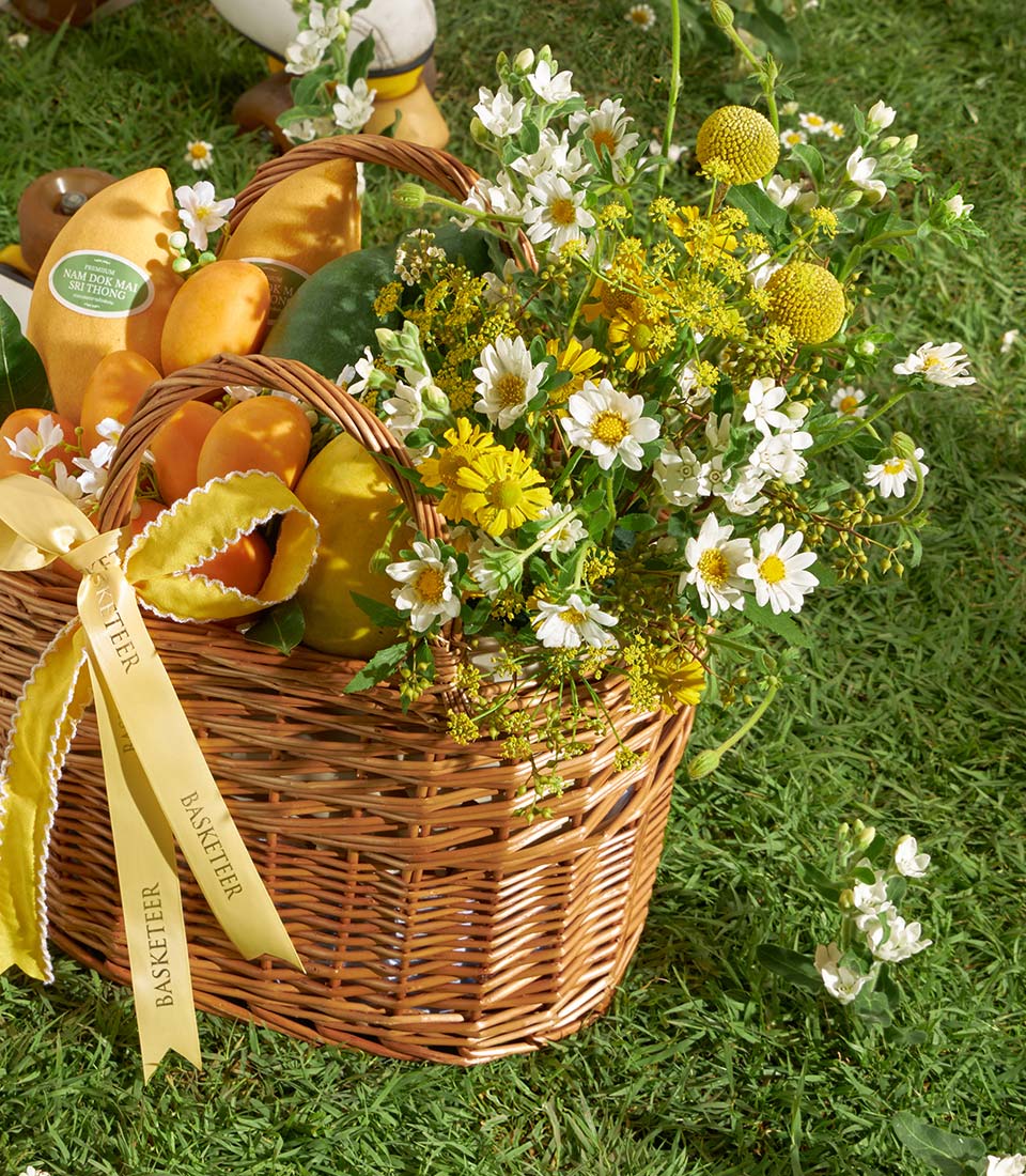 Wicker gift basket with Nam Dok Mai mangoes, white daisies, and yellow blooms on green grass