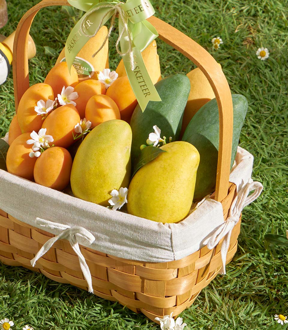 Woven basket filled with fresh Thai mangoes and white flowers, tied with green ribbon for premium gifting