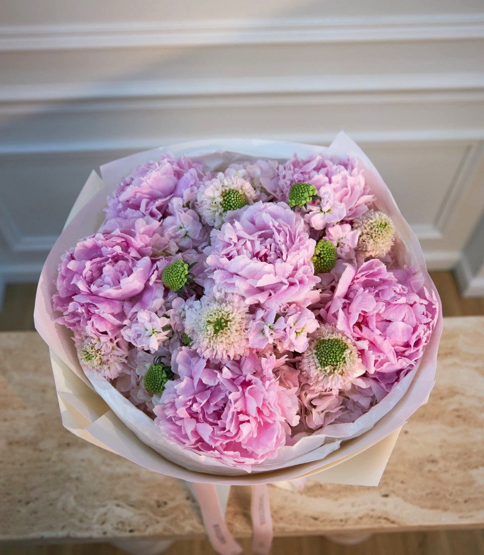 Pink and lavender peonies with green button mums wrapped in white paper with pink ribbon, displayed indoors on a marble countertop.