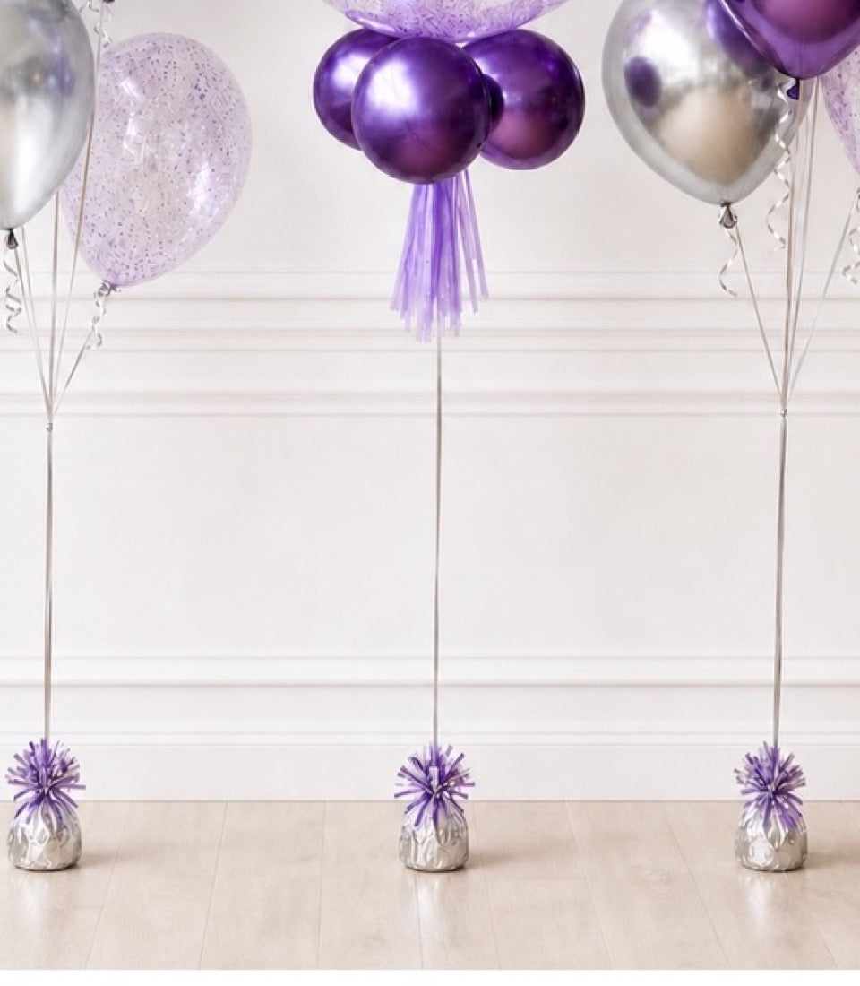 Purple and silver balloon bouquets with tulle tassels and weighted bases on light wood floor against white wall.