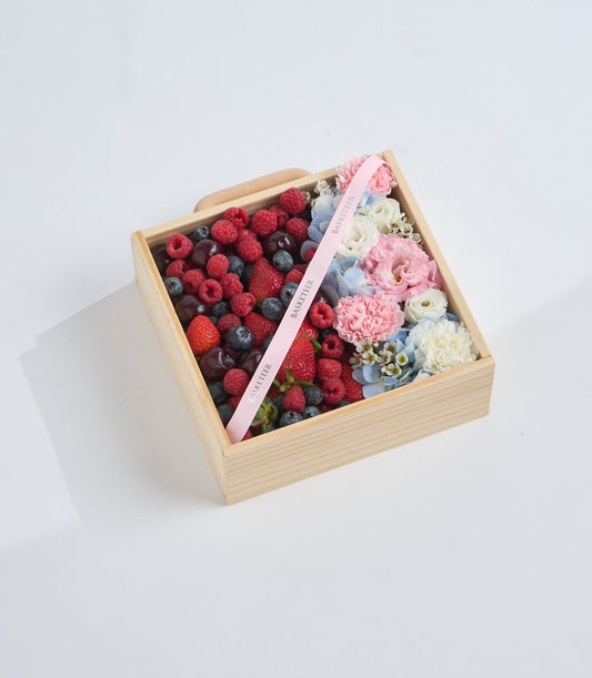 Wooden box gift arrangement with fresh raspberries, blueberries, and cherries on left side, pink peonies, white roses, and blue eucalyptus flowers on right with pink ribbon.