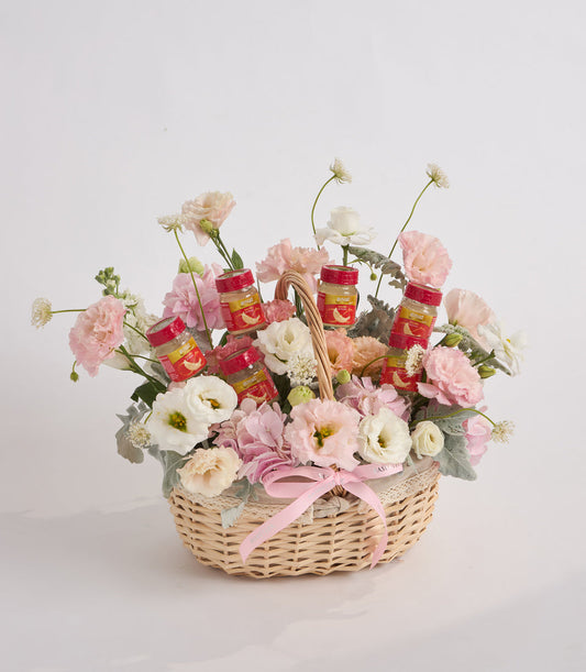 Wicker basket with pink and white carnations, roses, and daisies, accented with red jam jars and a pink ribbon bow.