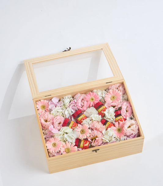 Wooden floral gift box with glass lid containing pink gerberas, white flowers, and red decorative items on white background.