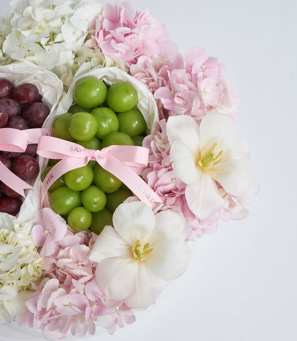 Luxury gift arrangement featuring pink and white peonies, white orchids, green and red grapes in tissue-wrapped containers with pink ribbon bows.