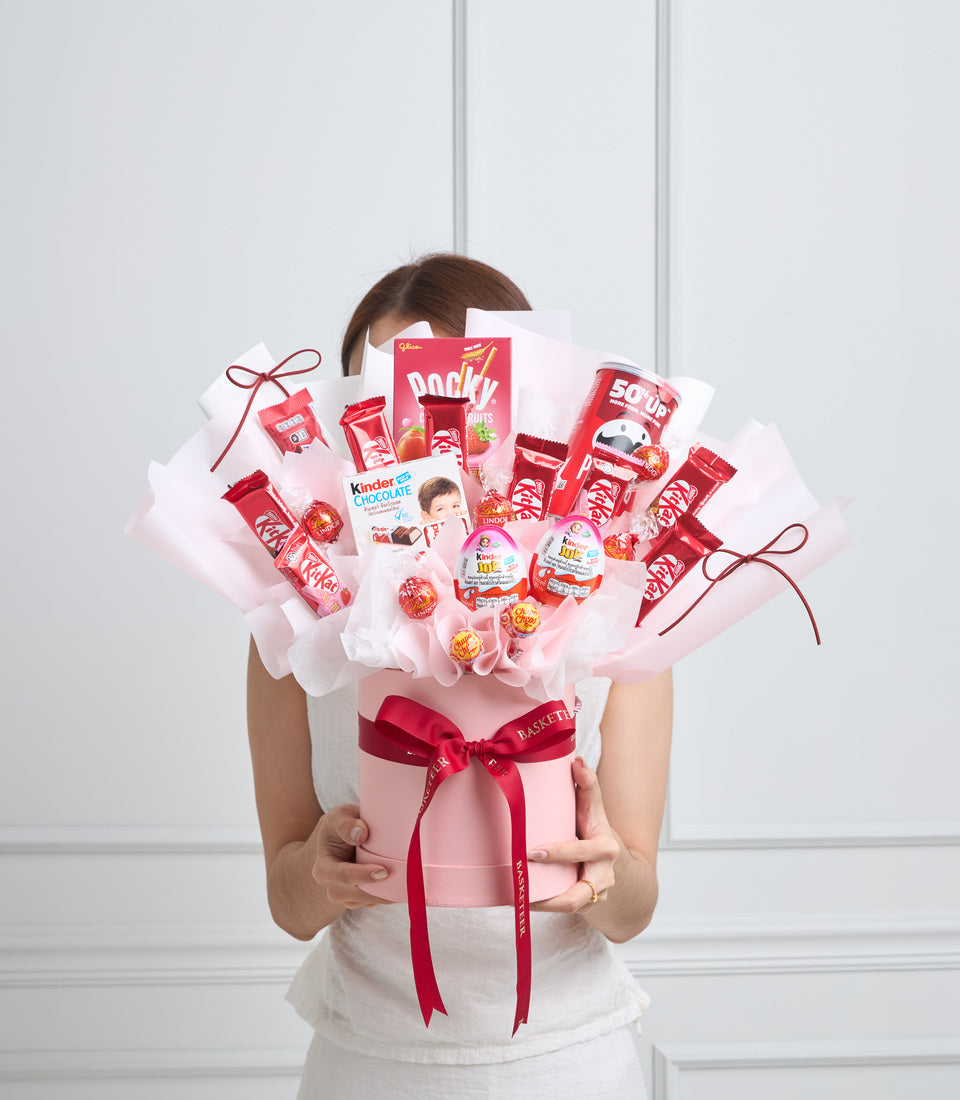 Pink candy bouquet with red ribbon bow held by woman, featuring Pocky, Kit Kat, Kinder Chocolate and lollipops in pink tissue wrapping.