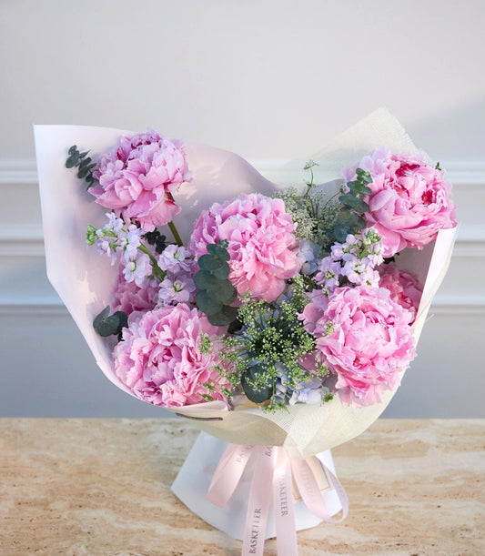 Luxurious bouquet of pink peonies with purple statice and baby's breath, wrapped in white paper with pink ribbons.
