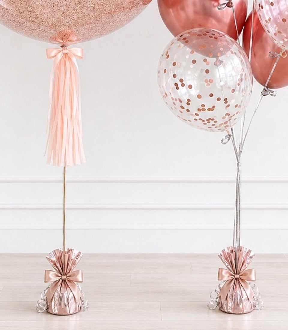 Rose gold balloons with confetti and tassels on weighted bases against white paneled wall backdrop.
